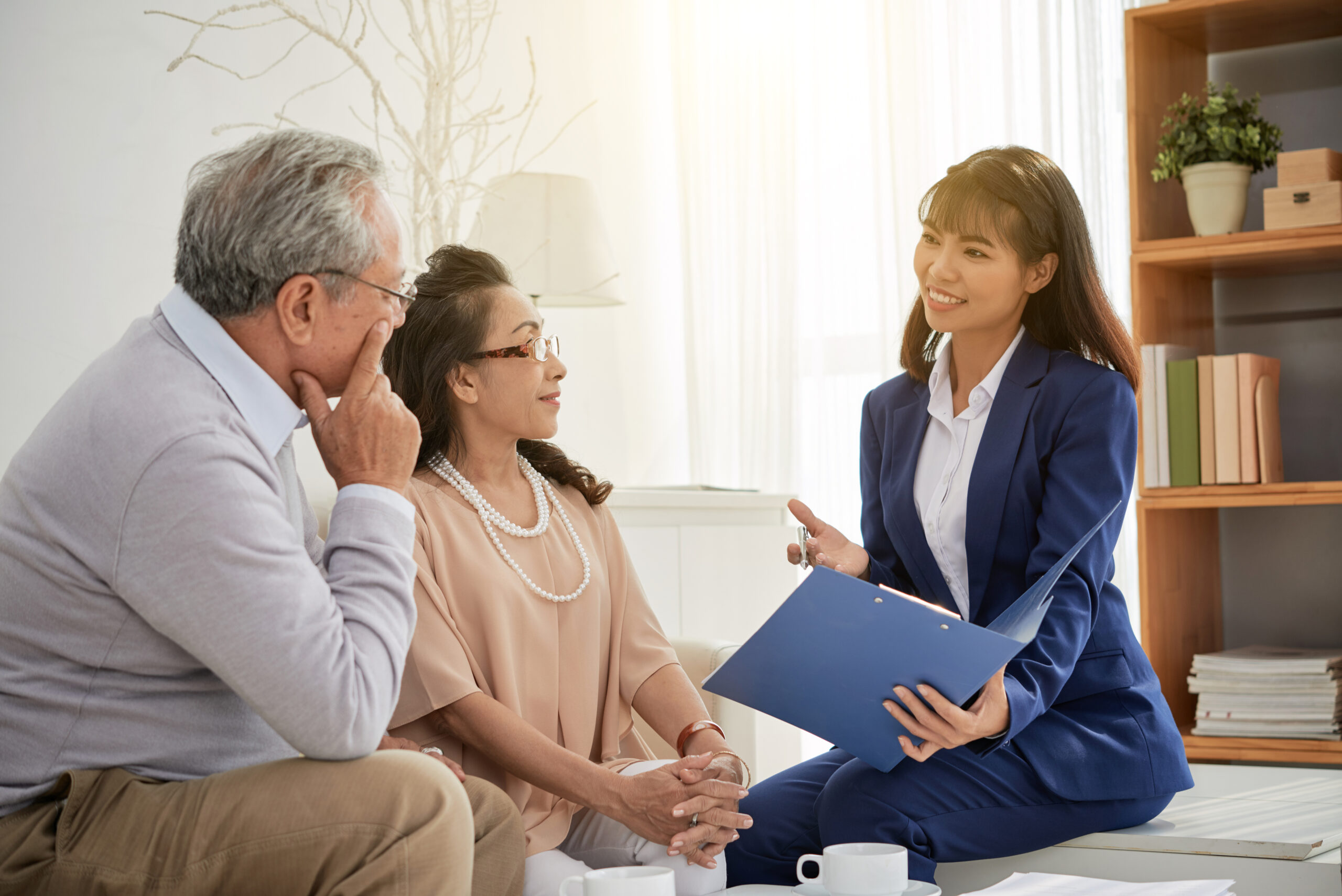 Attractive Asian estate agent with charming smile showing senior couple photos of apartments offered for sale while having meeting at cozy office